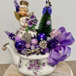 Puede incluir: Un arreglo decorativo en una tetera con una figura de ángel vintage con alas doradas, sosteniendo flores moradas. La tetera está llena de adornos morados, broches y un pequeño árbol de Navidad, acentuado con un gran lazo morado. La tetera tiene un diseño floral.