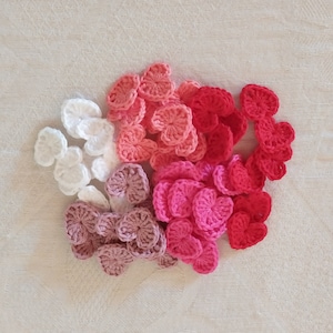 Puede incluir: Una colección de corazones de ganchillo en varios tonos de rojo, rosa y blanco. Los corazones hechos a mano están dispuestos sobre una superficie texturizada de color crema, creando una exhibición visualmente atractiva.