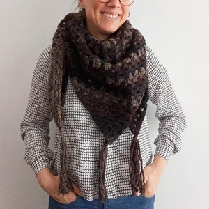 Puede incluir: Una mujer lleva una bufanda triangular de ganchillo marrón y negro con flecos. La bufanda se lleva alrededor del cuello y tiene un patrón texturizado.