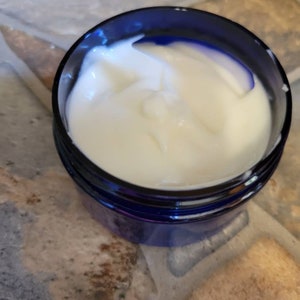 May include: A close-up of a blue jar filled with a white, creamy substance. The jar is sitting on a textured surface.