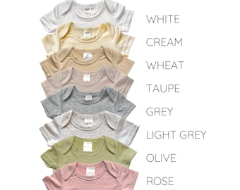 Könnte beinhalten: Ein Set aus acht Baby-Bodys in verschiedenen Farben, darunter Weiß, Creme, Weizen, Taupe, Grau, Hellgrau, Oliv und Rose. Die Bodys sind vertikal angeordnet, mit den Farbnamen rechts neben jedem Body. Der Text "Blanks by Tiny Wolfe" befindet sich oben im Bild.