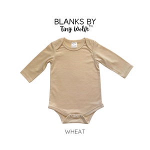 K&ouml;nnte beinhalten: Ein lang&auml;rmliger, beiger Baby-Body mit Rundhalsausschnitt und Druckkn&ouml;pfen. Der Body ist aus einem weichen, bequemen Stoff gefertigt und mit "Blanks by Tiny Wolfe" und "Wheat" beschriftet.