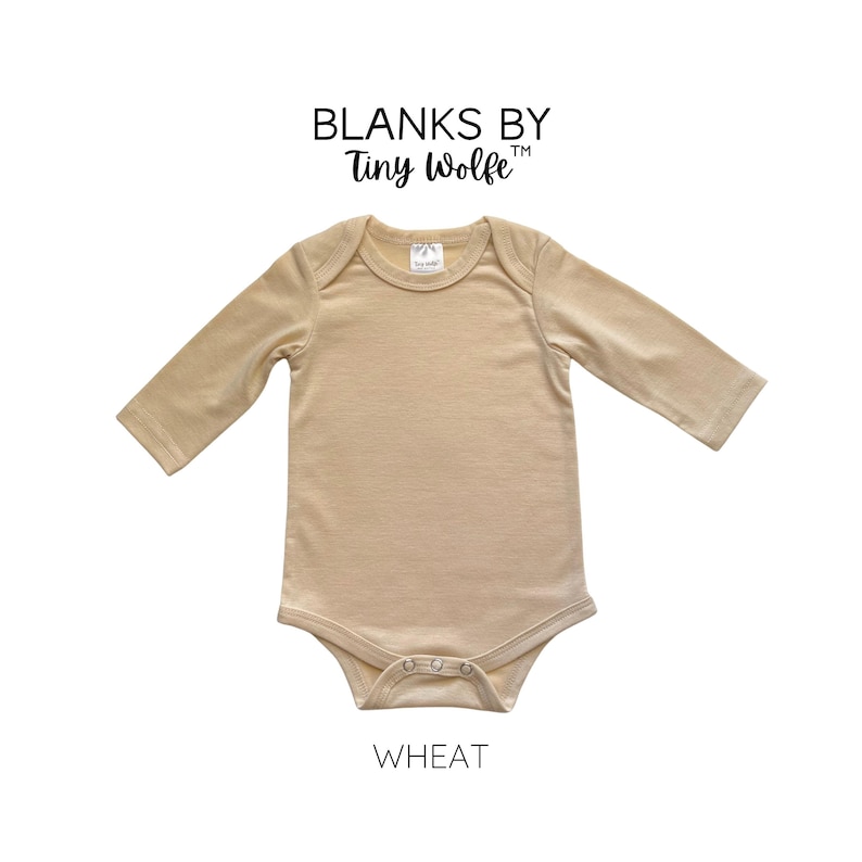 K&ouml;nnte beinhalten: Ein lang&auml;rmliger, beiger Baby-Body mit Rundhalsausschnitt und Druckkn&ouml;pfen. Der Body ist aus einem weichen, bequemen Stoff gefertigt und mit "Blanks by Tiny Wolfe" und "Wheat" beschriftet.