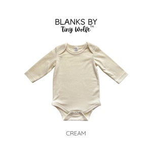 K&ouml;nnte beinhalten: Ein cremefarbener lang&auml;rmeliger Baby-Body mit Rundhalsausschnitt und Druckkn&ouml;pfen. Der Body ist aus weichem, bequemem Stoff gefertigt. Blanks by Tiny Wolfe ist oben auf dem Body gedruckt. Das Wort "CREAM" ist unter dem Body gedruckt.