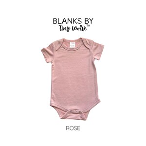 May include: A plain rose-colored baby onesie with short sleeves. The onesie has a snap closure at the bottom and is made of a soft, comfortable fabric. The text "Blanks by Tiny Wolfe" is printed above the onesie. The word "ROSE" is printed below the onesie.