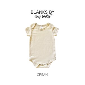 May include: A cream colored short-sleeved baby onesie with a round neckline. The onesie is made of a soft, comfortable fabric and has a snap closure at the bottom. The onesie is labeled "Blanks by Tiny Wolfe" and "Cream".
