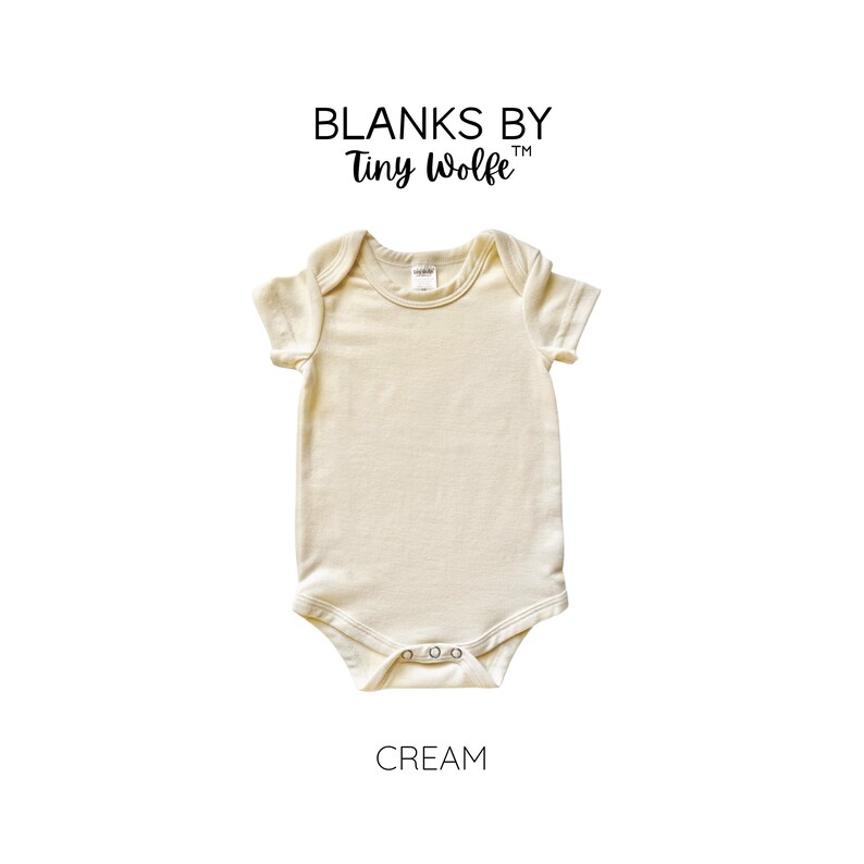 May include: A cream colored short-sleeved baby onesie with a round neckline. The onesie is made of a soft, comfortable fabric and has a snap closure at the bottom. The onesie is labeled "Blanks by Tiny Wolfe" and "Cream".
