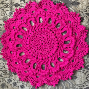 May include: A large, pink crocheted doily with a circular design. The doily has a centre circle with a raised texture and a repeating pattern of loops and bobbles.