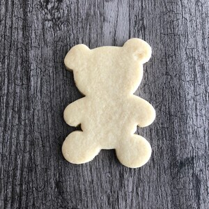 May include: A plain, un-decorated sugar cookie shaped like a teddy bear. The cookie is light brown and is sitting on a woodgrain surface.