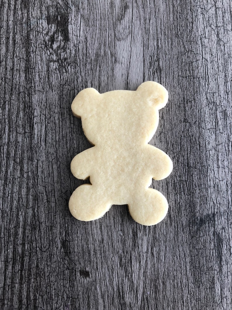 May include: A plain, un-decorated sugar cookie shaped like a teddy bear. The cookie is light brown and is sitting on a woodgrain surface.