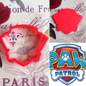 May include: Red plastic cookie cutter shaped like a dog's head. The cutter is on a floral patterned surface. A Paw Patrol logo is shown in the background.