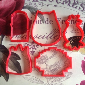 May include: Five red plastic cookie cutters in various shapes: a dress, a tulip, a cat, a seashell, and a decorative shape. The cutters are arranged on a floral background with text.