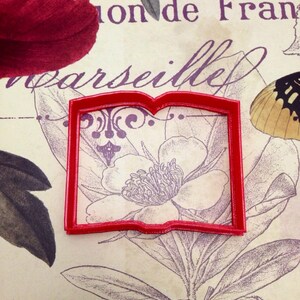 May include: Red plastic cookie cutter shaped like an open book. The cutter is on a vintage floral background with the text "ion de Fran" and "arseille".