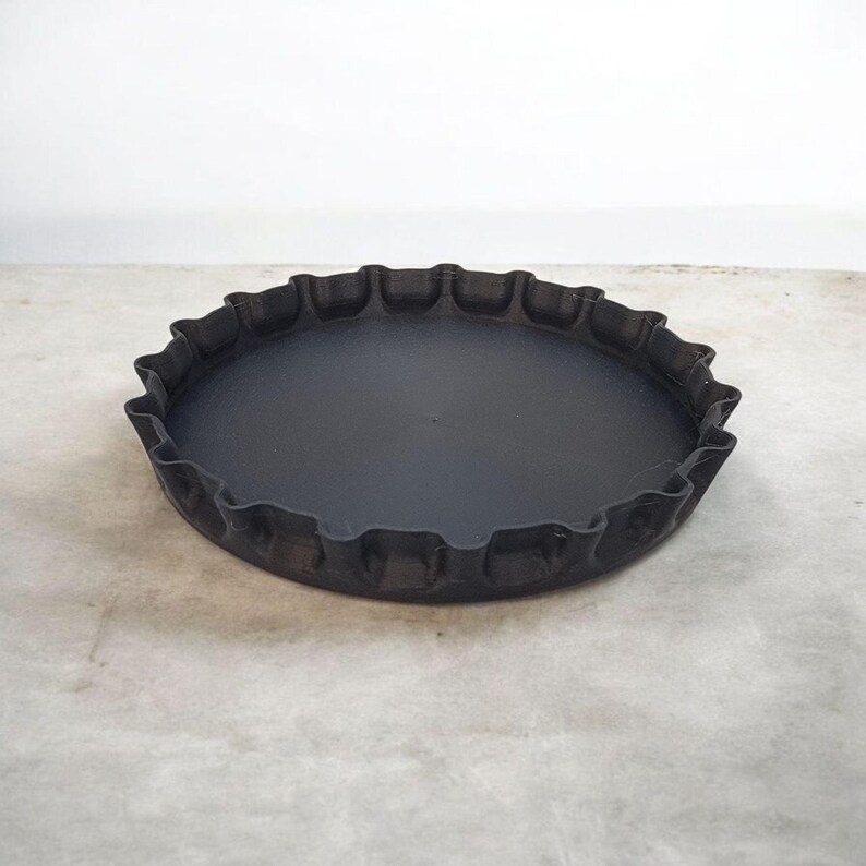 K&ouml;nnte beinhalten: Eine gro&szlig;e, mattschwarze Ablage in Form eines Flaschenverschlusses. Sie hat eine flache, kreisf&ouml;rmige Basis und einen markanten, gewellten Rand. Diese dekorative Ablage eignet sich zum Halten kleiner Gegenst&auml;nde.