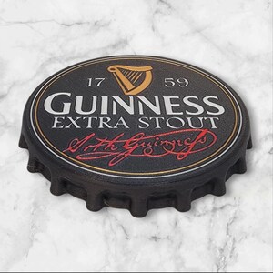 K&ouml;nnte beinhalten: Ein schwarzer Flaschenverschluss mit dem Guinness Extra Stout Logo. Das Logo zeigt eine goldene Harfe, den Schriftzug "GUINNESS EXTRA STOUT" in Wei&szlig; und die Unterschrift "Arth Guinness" in Rot. Der Verschluss hat eine strukturierte Oberfl&auml;che.