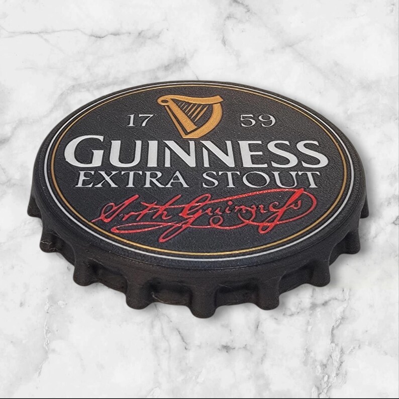 K&ouml;nnte beinhalten: Ein schwarzer Flaschenverschluss mit dem Guinness Extra Stout Logo. Das Logo zeigt eine goldene Harfe, den Schriftzug "GUINNESS EXTRA STOUT" in Wei&szlig; und die Unterschrift "Arth Guinness" in Rot. Der Verschluss hat eine strukturierte Oberfl&auml;che.