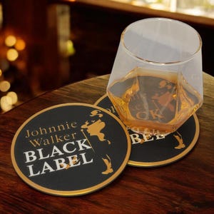K&ouml;nnte beinhalten: Ein klares, geometrisch geformtes Glas mit bernsteinfarbener Fl&uuml;ssigkeit steht auf einem schwarzen Untersetzer. Der Untersetzer tr&auml;gt die Aufschrift "Johnnie Walker BLACK LABEL" in wei&szlig;er und goldener Schrift. Ein weiterer Untersetzer ist sichtbar.