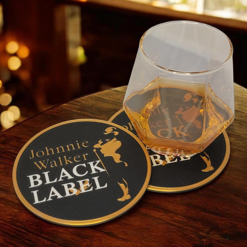 K&ouml;nnte beinhalten: Ein klares, geometrisch geformtes Glas mit bernsteinfarbener Fl&uuml;ssigkeit steht auf einem schwarzen Untersetzer. Der Untersetzer tr&auml;gt die Aufschrift "Johnnie Walker BLACK LABEL" in wei&szlig;er und goldener Schrift. Ein weiterer Untersetzer ist sichtbar.