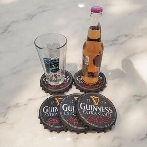 K&ouml;nnte beinhalten: Ein Bierglas und eine Flasche mit bernsteinfarbener Fl&uuml;ssigkeit stehen auf Untersetzern aus Guinness-Flaschenverschl&uuml;ssen. Die Untersetzer sind schwarz mit wei&szlig;em Text und einem Harfenlogo. Die Flasche hat einen lila Verschluss.