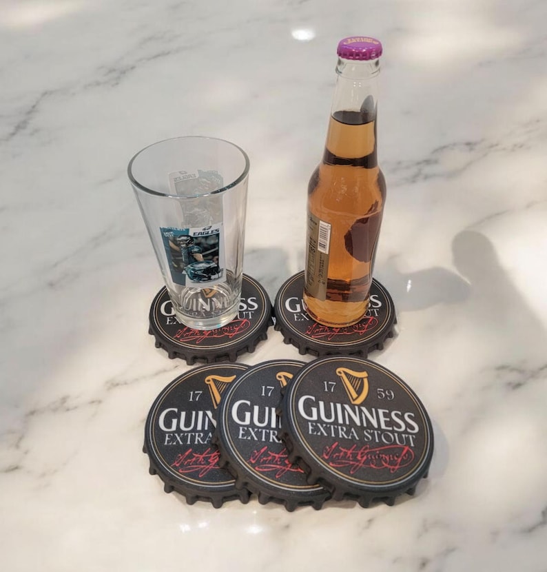 K&ouml;nnte beinhalten: Ein Bierglas und eine Flasche mit bernsteinfarbener Fl&uuml;ssigkeit stehen auf Untersetzern aus Guinness-Flaschenverschl&uuml;ssen. Die Untersetzer sind schwarz mit wei&szlig;em Text und einem Harfenlogo. Die Flasche hat einen lila Verschluss.