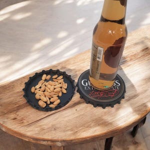 K&ouml;nnte beinhalten: Ein schwarzer Untersetzer in Form eines Flaschenverschlusses mit dem Text "Guinness Extra Stout" tr&auml;gt eine Bierflasche. Ein zweiter schwarzer Teller in Form eines Flaschenverschlusses enth&auml;lt Erdn&uuml;sse. Die Artikel liegen auf einem Holztisch.