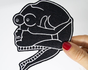 May include: Black embroidered patch of a creature with three eyes, a large mouth, and a long, thin body. The creature is outlined in white thread.