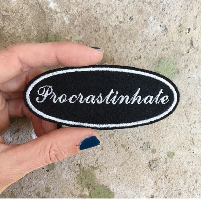 May include: Black oval patch with white embroidered text that reads "Procrastinate". The patch has a white border and is held by a hand against a textured background. The patch is a humorous accessory.