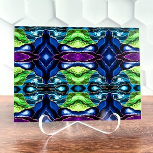 May include: A rectangular art piece with a symmetrical design in shades of blue, green, and purple, accented with gold lines. The artwork is displayed on a clear stand, set against a white geometric background.