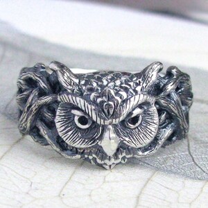 May include: A silver ring featuring an owl design. The owl's eyes are black and the ring is detailed with feathers.
