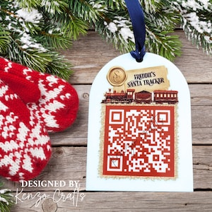 May include: A white arched ornament with a dark blue ribbon featuring a red steam train graphic, a gold wax seal, and the text Freddie's Santa Tracker above a large red and white QR code. The ornament is displayed against a rustic wooden background.