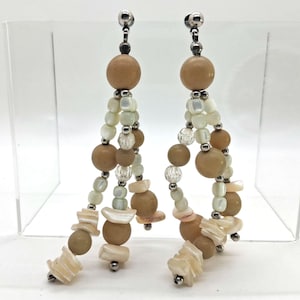 May include: A pair of dangle earrings featuring a combination of beige, white, and clear beads. The earrings have a silver-tone post and are composed of round beads, small white beads, and shell-shaped accents. The design is symmetrical and elegant.