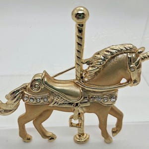 以下が含まれることがあります： 金色のユニコーンブローチ。メリーゴーランドの馬を連想させる。サドル、たてがみ、角など、細部までこだわったデザイン。サドルにはクリアなラインストーンがセットされている。ユニコーンは、装飾的なトップが付いた垂直のポールに取り付けられている。