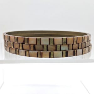 May include: A set of three stacked bangles featuring a repeating pattern of rectangular iridescent shell tiles alternating with thin vertical brass metal bars. The bangles have a smooth, circular profile with a warm, metallic and pearlescent finish.