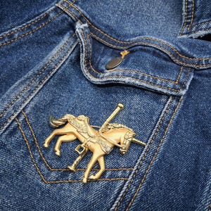 以下が含まれることがあります： 青いデニムジャケットのポケットに、メリーゴーランドのデザインの金色のユニコーンブローチがピン留めされています。ブローチは、青い生地とコントラストの効いた黄色のステッチに対して、サドルとポールを備えたユニコーンが特徴です。
