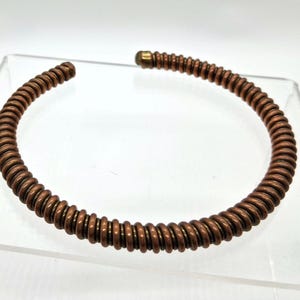 May include: A copper-colored, open-ended cuff bracelet. The bracelet is made of a series of tightly wound, ridged metal rings. The ends of the bracelet are smooth, with a small, gold-colored cap on one end. The bracelet is displayed on a clear surface.