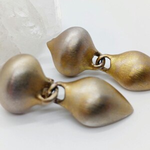 May include: A pair of gold-toned earrings with a teardrop shape. The earrings have a brushed finish and a subtle two-tone effect.