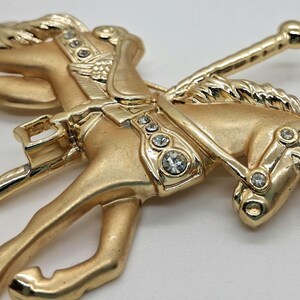以下が含まれることがあります： 透明なラインストーンがアクセントになった、金色のメリーゴーランドの馬のブローチ。馬は走っている姿で描かれ、鞍とたてがみが細かく表現されています。クラシックでヴィンテージな雰囲気のブローチです。