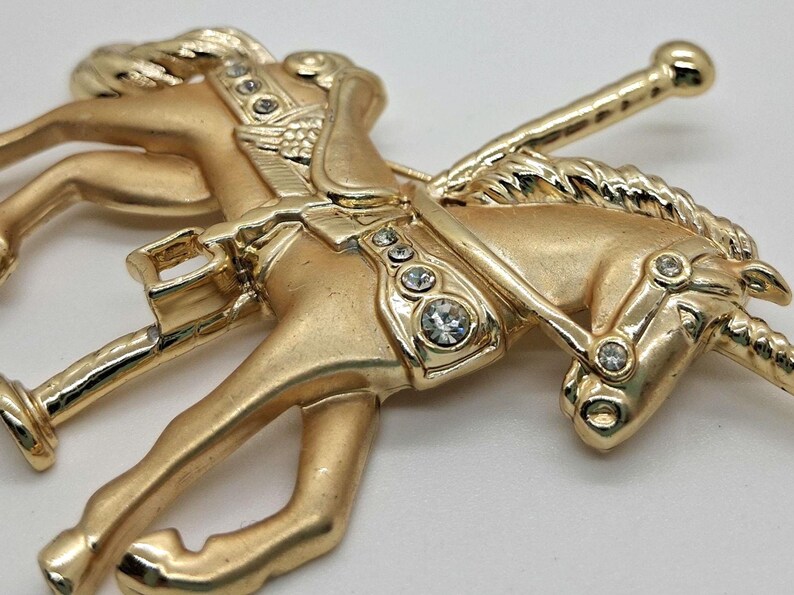 以下が含まれることがあります： 透明なラインストーンがアクセントになった、金色のメリーゴーランドの馬のブローチ。馬は走っている姿で描かれ、鞍とたてがみが細かく表現されています。クラシックでヴィンテージな雰囲気のブローチです。