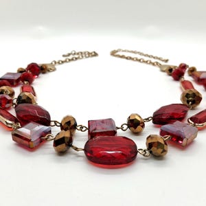 May include: A two-strand necklace featuring faceted red and bronze-colored beads. The necklace has a gold-colored chain and clasp. The beads are various shapes, including squares, rectangles, and round shapes.