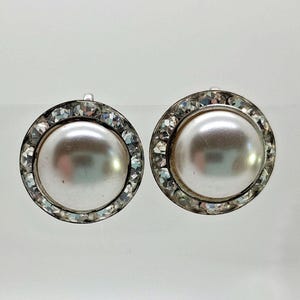 May include: A pair of round, silver-toned earrings. Each earring features a large, lustrous, off-white pearl center, surrounded by a halo of sparkling, clear crystals. The earrings are set against a plain, light background.