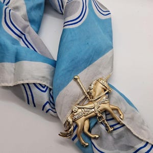 以下が含まれることがあります： 透明なクリスタルアクセントが付いた金色のメリーゴーランドの馬のブローチが、青、白、グレーの模様の入ったスカーフにピン留めされています。 スカーフは折りたたまれ、ドレープされており、そのデザインが示されています。 ブローチは詳細なデザインが特徴です。