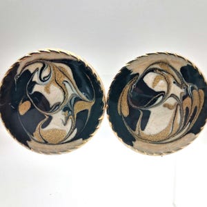 May include: Pair of round earrings with a swirling pattern in black, gold, and cream. The earrings have a textured, dark border. The design features a marbled effect, creating a unique and artistic look. These earrings are suitable for everyday wear or special occasions.