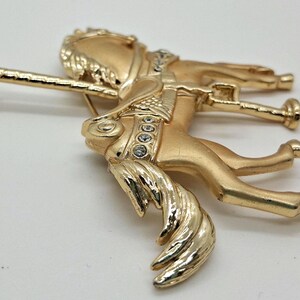 以下が含まれることがあります： 詳細なデザインの金色のメリーゴーランドの馬のブローチ。ブローチは、たてがみと尾が流れる、ギャロップのポーズの馬が特徴です。馬は透明なクリスタルのアクセントと装飾的なサドルで飾られています。ブローチの裏側にはピン留めがあります。