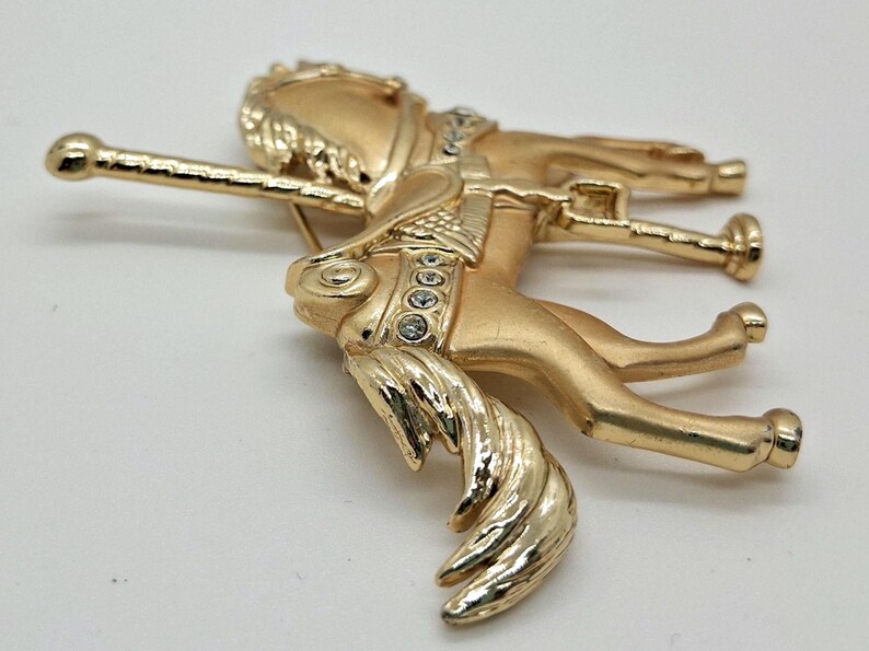 以下が含まれることがあります： 詳細なデザインの金色のメリーゴーランドの馬のブローチ。ブローチは、たてがみと尾が流れる、ギャロップのポーズの馬が特徴です。馬は透明なクリスタルのアクセントと装飾的なサドルで飾られています。ブローチの裏側にはピン留めがあります。