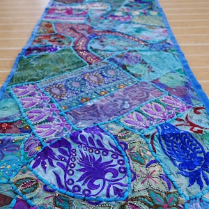 May include: A blue patchwork table runner with colorful embroidery and sequins. The runner features a variety of floral and paisley designs.