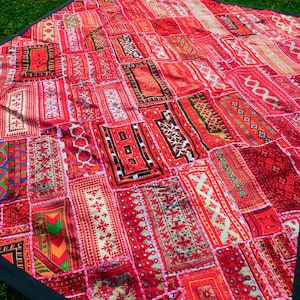 May include: A vibrant red patchwork quilt with a black border. The quilt is composed of numerous rectangular patches, each featuring intricate patterns in shades of red, pink, and orange. The quilt is laid out on green grass.