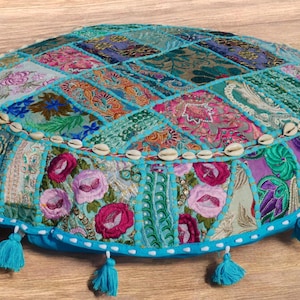 Könnte beinhalten: Ein rundes türkisfarbenes Bodenkissen mit Patchwork-Muster aus bunten Stoffen und gestickten Blumen. Das Kissen ist mit Weiß eingefasst und hat eine Reihe weißer Muscheln am Rand. Es hat türkisfarbene Quasten, die von unten hängen.