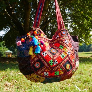 Könnte beinhalten: Eine große, farbenfrohe Patchwork-Tragetasche mit Bohème-Design. Die Tasche hat aufwendige Stickereien in Rot, Orange, Grün und Blau mit dekorativen Quasten und Perlenakzenten. Die Tasche hängt an einem roten Riemen.