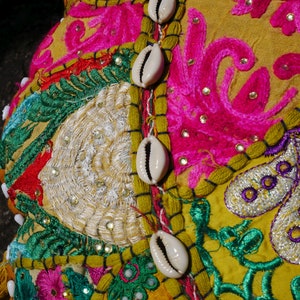 May include: A yellow fabric cushion featuring colourful embroidery and white cowrie shells. The cushion is adorned with floral patterns and intricate details.