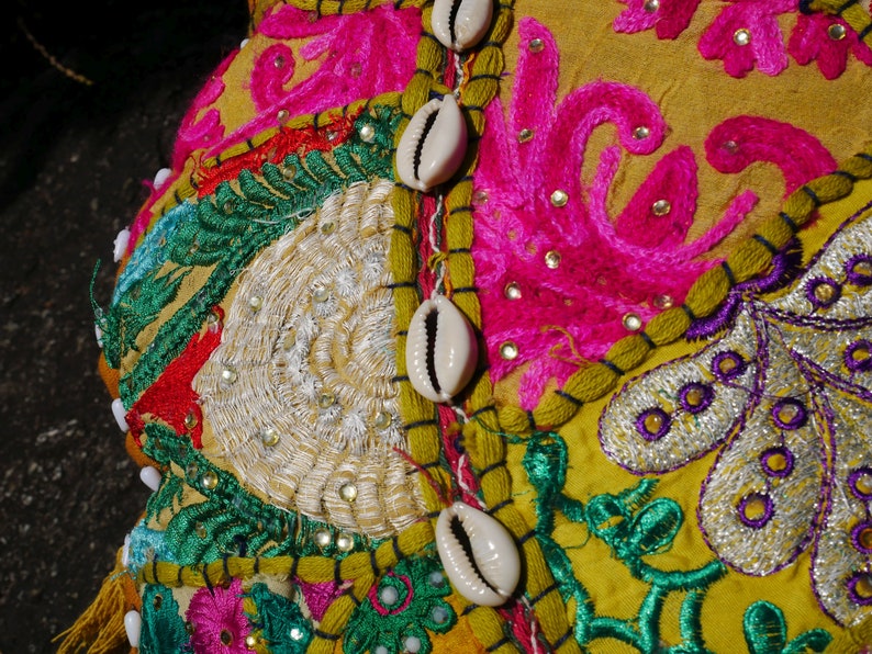 May include: A yellow fabric cushion featuring colourful embroidery and white cowrie shells. The cushion is adorned with floral patterns and intricate details.
